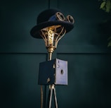 A vintage-style lamp incorporates a top hat and goggles, mounted on a brass frame with an exposed Edison bulb. It stands on a tripod base with a box-like feature in a dark teal setting.