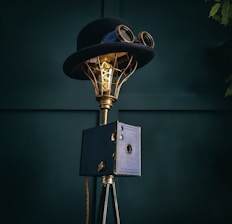 A vintage-style lamp incorporates a top hat and goggles, mounted on a brass frame with an exposed Edison bulb. It stands on a tripod base with a box-like feature in a dark teal setting.