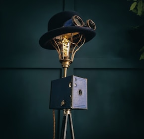 A vintage-style lamp incorporates a top hat and goggles, mounted on a brass frame with an exposed Edison bulb. It stands on a tripod base with a box-like feature in a dark teal setting.