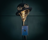 A creatively designed lamp with a vintage camera base, topped with a wire-frame light bulb wearing a classic bowler hat and goggles. The setup merges elements of imagery and whimsy in a steampunk style.