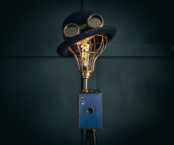 A creatively designed lamp with a vintage camera base, topped with a wire-frame light bulb wearing a classic bowler hat and goggles. The setup merges elements of imagery and whimsy in a steampunk style.