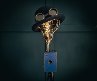 A creatively designed lamp with a vintage camera base, topped with a wire-frame light bulb wearing a classic bowler hat and goggles. The setup merges elements of imagery and whimsy in a steampunk style.