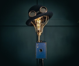 A creatively designed lamp with a vintage camera base, topped with a wire-frame light bulb wearing a classic bowler hat and goggles. The setup merges elements of imagery and whimsy in a steampunk style.