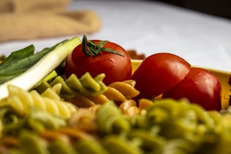 A close-up of a colorful assortment of uncooked fusilli pasta, including green, white, and yellow varieties, accompanied by fresh red tomatoes and green vegetables such as leeks.