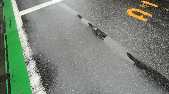 A wet road surface with visible white and green lane markings. The road appears freshly wet, likely due to rain, with reflections visible on the asphalt. There is a partial yellow marking or letter on the right-hand side.