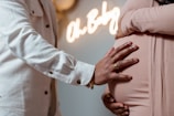 A close-up of two people embracing, with one hand resting on the pregnant belly of the other. The background includes a softly focused neon sign reading 'Oh Baby.' The scene suggests a warm and intimate moment.