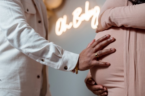 A close-up of two people embracing, with one hand resting on the pregnant belly of the other. The background includes a softly focused neon sign reading 'Oh Baby.' The scene suggests a warm and intimate moment.