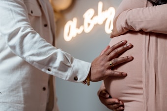 A close-up of two people embracing, with one hand resting on the pregnant belly of the other. The background includes a softly focused neon sign reading 'Oh Baby.' The scene suggests a warm and intimate moment.