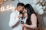 A couple stands close together, holding ultrasound images. The woman is wearing a blush-colored dress, and the man is in a white shirt. In the background, there are white and metallic balloons, and a neon sign reads 'Oh Baby'.