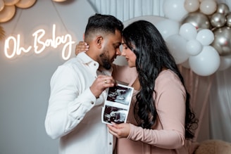 A couple stands close together, holding ultrasound images. The woman is wearing a blush-colored dress, and the man is in a white shirt. In the background, there are white and metallic balloons, and a neon sign reads 'Oh Baby'.
