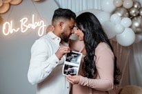 A couple stands close together, holding ultrasound images. The woman is wearing a blush-colored dress, and the man is in a white shirt. In the background, there are white and metallic balloons, and a neon sign reads 'Oh Baby'.