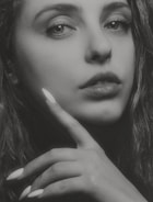 A close-up black and white photograph of a woman's face, focusing on her eyes and lips. Her hand is gently touching her chin with her fingers, displaying long, manicured nails. The lighting creates a contrast, emphasizing the texture of her skin and hair.
