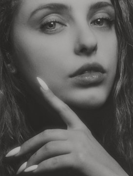 A close-up black and white photograph of a woman's face, focusing on her eyes and lips. Her hand is gently touching her chin with her fingers, displaying long, manicured nails. The lighting creates a contrast, emphasizing the texture of her skin and hair.