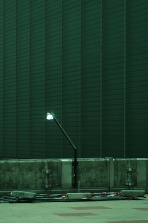 A dimly lit street lamp is positioned against a background of tall, dark green building panels. Below the lamp, there is a concrete barrier with visible metal fixtures and scattered cables on the ground.