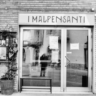 A black and white photograph of a storefront with glass doors reflecting the street scene across from it. The name 'I Malpensanti' is written above the entrance. Inside the window reflection, parts of a building and a person's silhouette holding a camera can be seen. On the left side, there is a small wall-mounted display with various photos and flyers, and a potted plant sits beneath it.