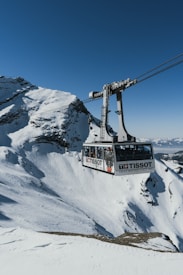 A cable car suspended over a snowy, mountainous landscape with clear blue skies. The cable car is branded with Tissot logos and is filled with passengers. The rugged, snow-covered mountains create a serene and adventurous atmosphere.