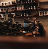A cozy coffee shop scene showing a barista making coffee with an espresso machine. Shelves in the background are stocked with coffee bags. The atmosphere is warm and inviting, with low lighting.