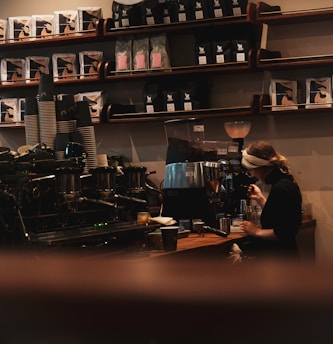 A cozy coffee shop scene showing a barista making coffee with an espresso machine. Shelves in the background are stocked with coffee bags. The atmosphere is warm and inviting, with low lighting.