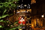 A festive city scene featuring a well-lit building adorned with holiday decorations and a prominent wreath. The foreground includes lush green trees with sparkling lights, enhancing the holiday spirit. The building is illuminated with an array of warm lights and ornate architectural details.