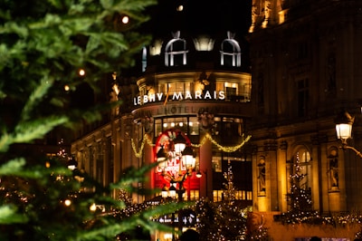 A festive city scene featuring a well-lit building adorned with holiday decorations and a prominent wreath. The foreground includes lush green trees with sparkling lights, enhancing the holiday spirit. The building is illuminated with an array of warm lights and ornate architectural details.