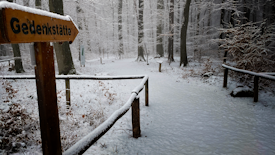 A snowy forest scene with tall trees covered in snow. In the foreground, there is a wooden sign with the word 'Gedenkst&auml;tte' and a snow-covered path leading deeper into the forest. The atmosphere is quiet and serene, with a light mist in the background, adding a sense of calm and solitude.