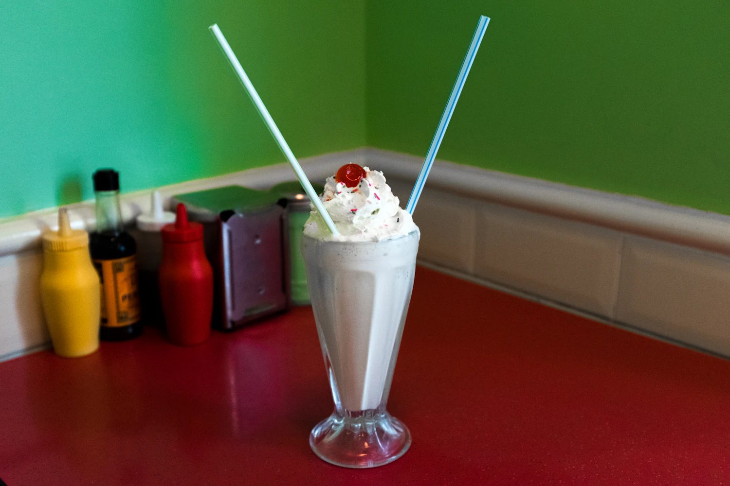 Vanilla cream soda topped with whipped cream and a cherry, two striped straws, on a red counter against a green wall