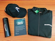 A black baseball cap, a black zip-up jacket, a black thermos, and a greeting card are arranged neatly on a wooden surface. The cap and jacket both display the word 'ASCENDION'. The greeting card reads 2023, includes a thank you message, and features a design with fireworks.