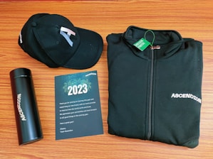 A black baseball cap, a black zip-up jacket, a black thermos, and a greeting card are arranged neatly on a wooden surface. The cap and jacket both display the word 'ASCENDION'. The greeting card reads 2023, includes a thank you message, and features a design with fireworks.