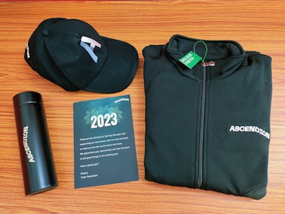 A black baseball cap, a black zip-up jacket, a black thermos, and a greeting card are arranged neatly on a wooden surface. The cap and jacket both display the word 'ASCENDION'. The greeting card reads 2023, includes a thank you message, and features a design with fireworks.