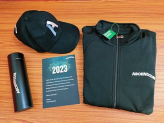 A black baseball cap, a black zip-up jacket, a black thermos, and a greeting card are arranged neatly on a wooden surface. The cap and jacket both display the word 'ASCENDION'. The greeting card reads 2023, includes a thank you message, and features a design with fireworks.