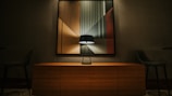 A modern interior scene featuring a large abstract painting with geometric shapes and muted colors. A sleek black table lamp is placed on top of a wooden sideboard, casting subtle light and shadows on the artwork. The room includes two dark upholstered chairs on either side, creating a balanced composition.