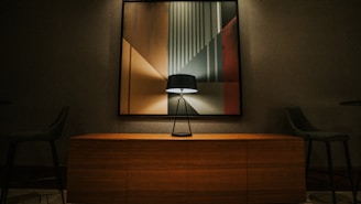 A modern interior scene featuring a large abstract painting with geometric shapes and muted colors. A sleek black table lamp is placed on top of a wooden sideboard, casting subtle light and shadows on the artwork. The room includes two dark upholstered chairs on either side, creating a balanced composition.
