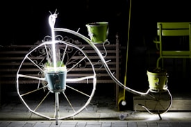 A decorative metal bicycle-shaped plant stand is illuminated with bright white lights. It holds three plant pots, with one at the center of the large front wheel and two on the handlebars, each containing plants. The scene is set against a dark background, with a wooden bench and a green chair partially visible.