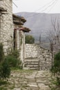 A rustic stone pathway leads to a quaint stone building with a red sign indicating a guesthouse. The setting features old stone walls and lush greenery on either side of the path. In the background, a mountainous landscape under a cloudy sky completes the scene, imparting a sense of tranquility.
