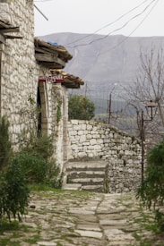 A rustic stone pathway leads to a quaint stone building with a red sign indicating a guesthouse. The setting features old stone walls and lush greenery on either side of the path. In the background, a mountainous landscape under a cloudy sky completes the scene, imparting a sense of tranquility.