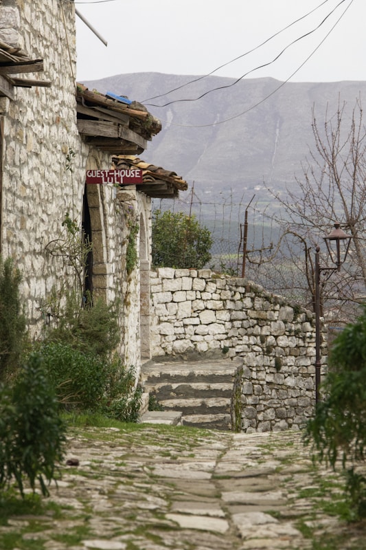 A rustic stone pathway leads to a quaint stone building with a red sign indicating a guesthouse. The setting features old stone walls and lush greenery on either side of the path. In the background, a mountainous landscape under a cloudy sky completes the scene, imparting a sense of tranquility.