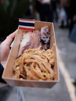 A close-up of a hand holding a cardboard container filled with grilled meat and noodles, adorned with a small flag resembling the Thai national flag.