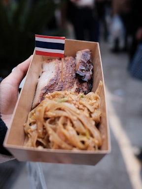A close-up of a hand holding a cardboard container filled with grilled meat and noodles, adorned with a small flag resembling the Thai national flag.