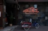 A vintage-style bicycle with a red metal basket is parked in front of a dark tiled wall. Above, a neon sign reads 'La Maison Lyonnaise' with silhouettes of kitchenware. A tall potted plant is placed beside the bicycle on a wooden platform.