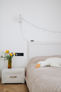 A cozy bedroom with a neatly made bed, featuring a light beige quilt. A stuffed white duck with an orange beak rests on the bed. Next to the bed, there is a white nightstand with a decorative yellow vase containing fresh yellow flowers. A string of delicate fairy lights is hanging on the wall above the nightstand.