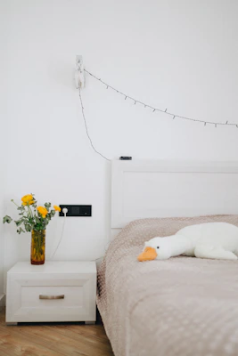 A cozy bedroom with a neatly made bed, featuring a light beige quilt. A stuffed white duck with an orange beak rests on the bed. Next to the bed, there is a white nightstand with a decorative yellow vase containing fresh yellow flowers. A string of delicate fairy lights is hanging on the wall above the nightstand.