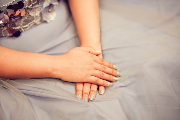 Hands are resting gently on a soft, light blue fabric with a decorative sequin pattern near the top. The nails are painted a light shade with sparkling accents.