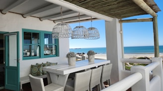 A coastal patio features a long white dining table surrounded by woven chairs. Above the table hang three rattan light fixtures. The patio is sheltered by a wooden pergola and overlooks a sandy beach and clear blue sea. The setting is decorated with teal window frames and cushions with intricate patterns, adding a touch of color to the otherwise neutral palette.