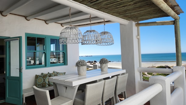 A coastal patio features a long white dining table surrounded by woven chairs. Above the table hang three rattan light fixtures. The patio is sheltered by a wooden pergola and overlooks a sandy beach and clear blue sea. The setting is decorated with teal window frames and cushions with intricate patterns, adding a touch of color to the otherwise neutral palette.