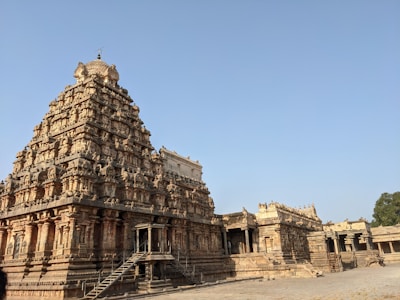 An intricately carved stone temple with a towering gopuram, showcasing traditional Indian architectural elements such as detailed sculptures and multiple tiers. The structure is surrounded by a paved courtyard under a clear blue sky.