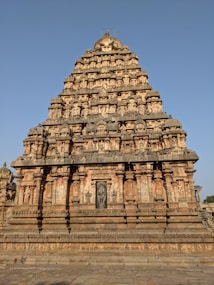A detailed view of a historical temple with intricate carvings and a pyramid-like structure. The architectural style is traditional, with numerous sculpted figures and ornamental details. The temple is set against a clear blue sky, highlighting its grandeur.