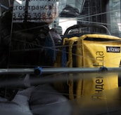 A person is sitting inside a bus or a public transport vehicle, holding a sign with partially visible text. Next to them is a large yellow bag, potentially used for deliveries, with some Cyrillic text and the number A12307 visible. The reflective glass creates an effect of lines crisscrossing over the scene.