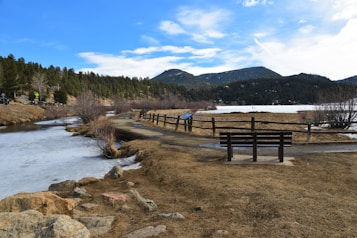 A serene lakeside scene surrounded by mountains with a path leading through dried grass towards the water. A solitary bench is positioned beside the path, inviting rest and reflection. The lake appears partially frozen, and the sky above is bright with a few scattered clouds.
