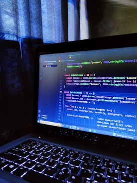 A laptop with illuminated keyboard keys, displaying a coding interface on its screen. The code includes JavaScript syntax highlighting with a visible sidebar showing a file tree. The window is dimly lit with curtains providing soft light in the background, suggesting a nighttime or indoor setting.