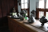 A collection of metal kitchenware, including various pots and lids, is arranged on a wooden kitchen counter. The room features wooden walls and windows with light curtains, allowing natural sunlight to illuminate the space.